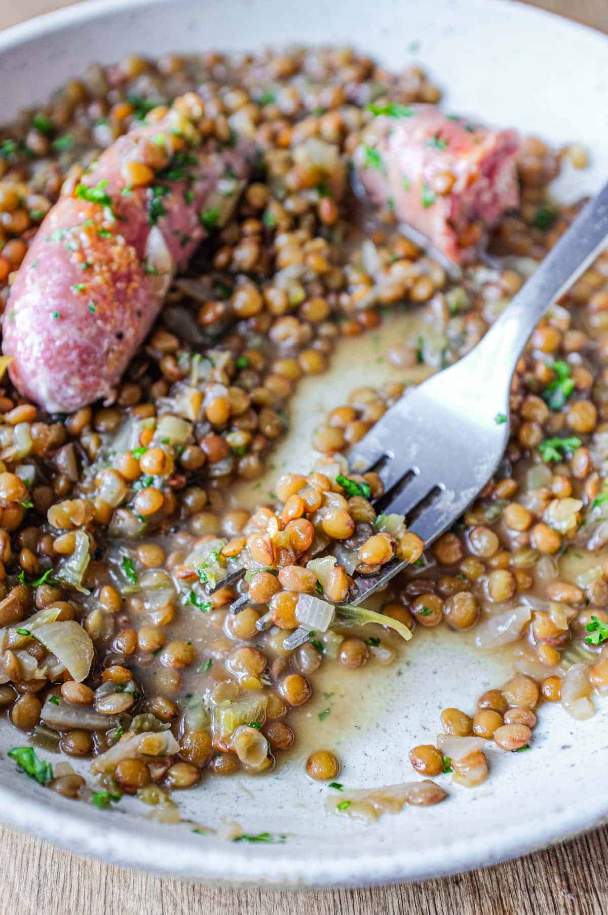 A shallow bowl has a layer of creamy lentils, topped with two Italian pork and fennel sausages. The dish is garnished with fresh parsley.