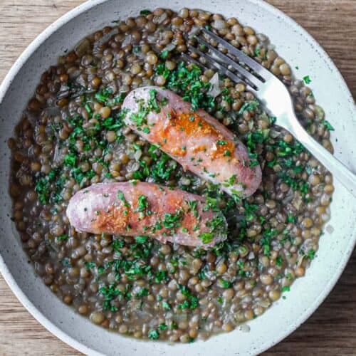 A shallow bowl has a layer of creamy lentils, topped with two Italian pork and fennel sausages. The dish is garnished with fresh parsley.