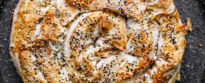 A coiled Turkish Börek on a baking sheet, scattered with sesame and nigella seeds.