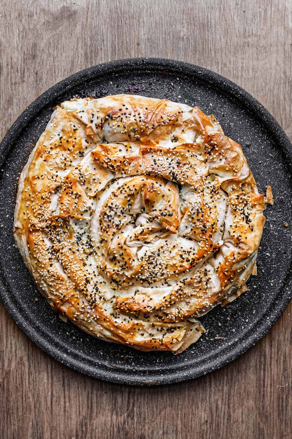 A coiled Turkish Börek on a baking sheet, scattered with sesame and nigella seeds.