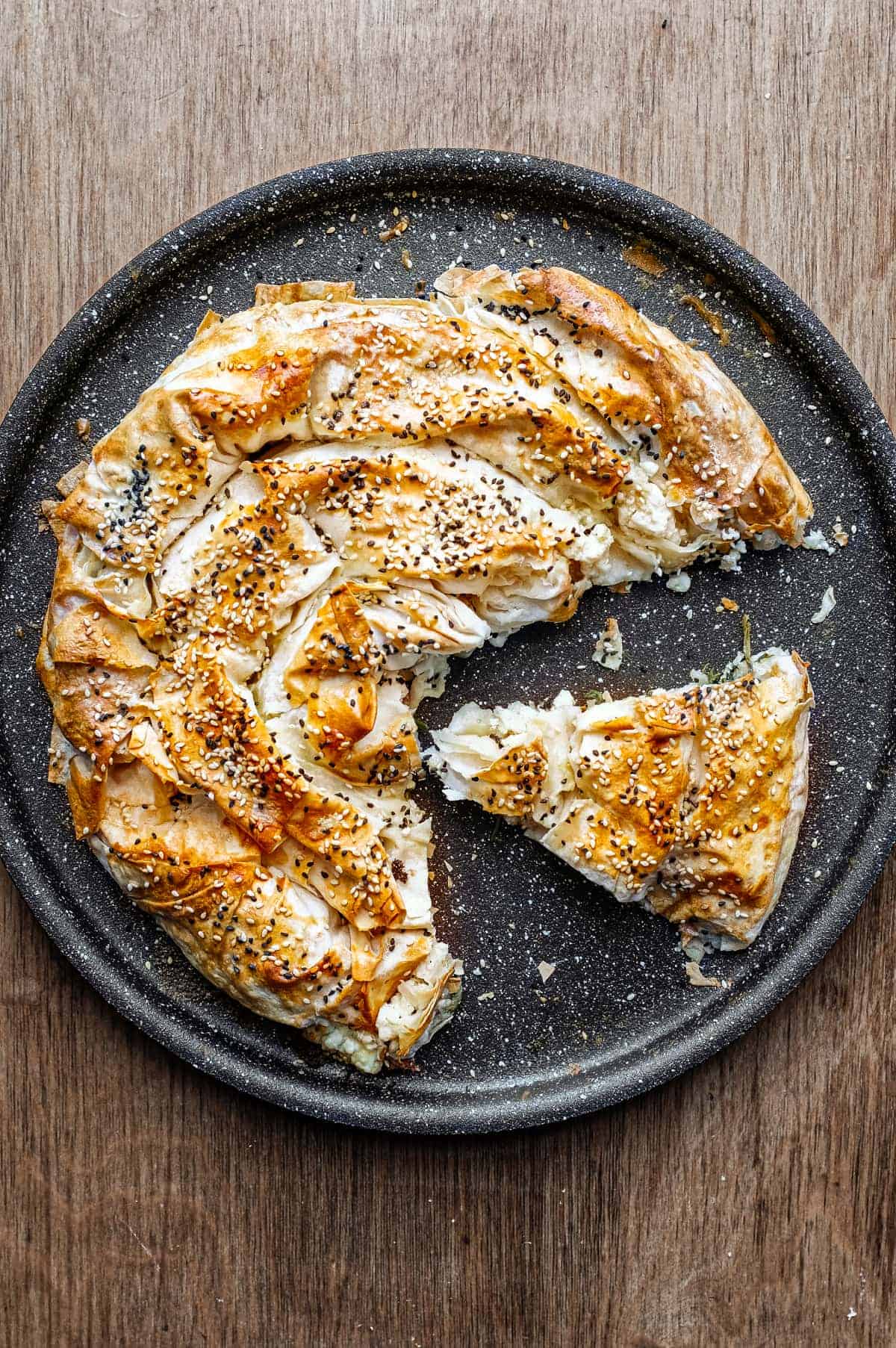 A  slice of a coiled Turkish Börek on a baking sheet, scattered with sesame and nigella seeds.