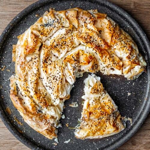 A  slice of a coiled Turkish Börek on a baking sheet, scattered with sesame and nigella seeds.