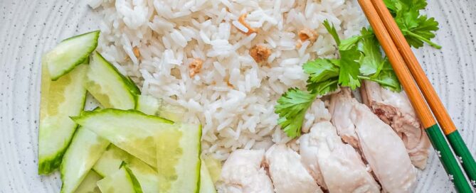 A plate of Hainan Chicken Rice with chopsticks. Featuring poached chicken, rice with specks of chicken skin, chopped cucumber and a sprig of cilantro. A small bowl of soup and an asian spoon sits to the side along with two condiments of chilli sauce and a ginger scallion sauce.