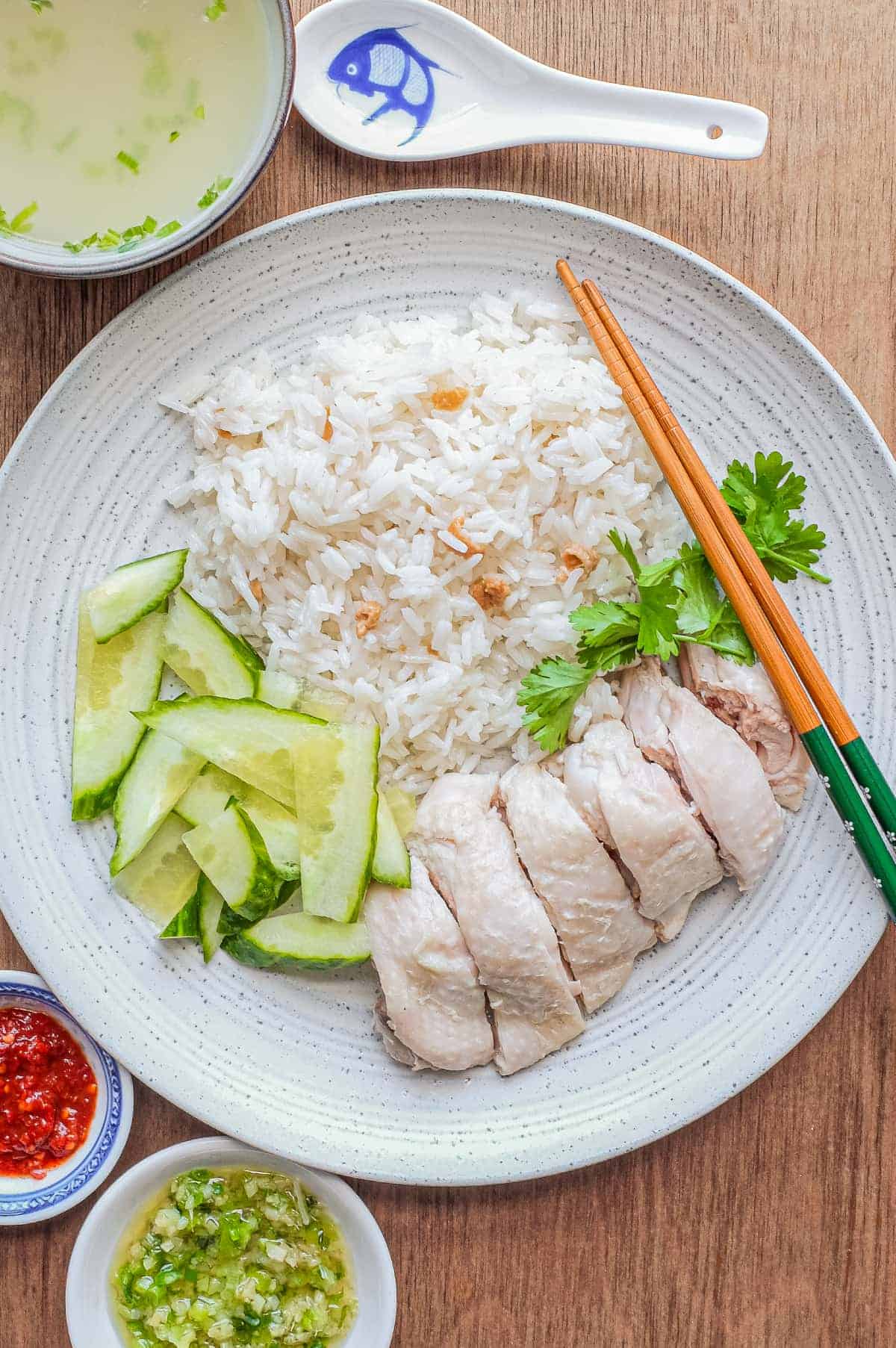 A plate of Hainan Chicken Rice with chopsticks. Featuring poached chicken, rice with specks of chicken skin, chopped cucumber and a sprig of cilantro. A small bowl of soup and an asian spoon sits to the side along with two condiments of chilli sauce and a ginger scallion sauce.