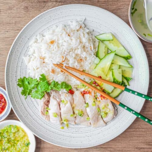 A plate of Hainan Chicken Rice with chopsticks. Featuring poached chicken, rice with specks of chicken skin, chopped cucumber and a sprig of cilantro. A small bowl of soup and an asian spoon sits to the side along with two condiments of chilli sauce and a ginger scallion sauce.