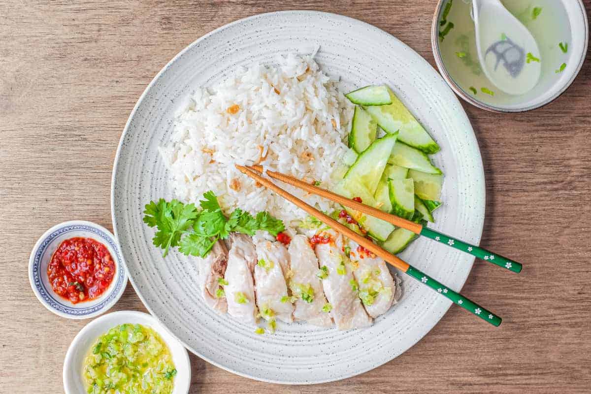 A plate of Hainan Chicken Rice with chopsticks. Featuring poached chicken, rice with specks of chicken skin, chopped cucumber and a sprig of cilantro. A small bowl of soup and an asian spoon sits to the side along with two condiments of chilli sauce and a ginger scallion sauce.