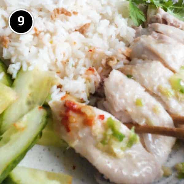 A plate of Hainan Chicken Rice with chopsticks. Featuring poached chicken, rice with specks of chicken skin, chopped cucumber and a sprig of cilantro. A small bowl of soup and an asian spoon sits to the side along with two condiments of chilli sauce and a ginger scallion sauce.