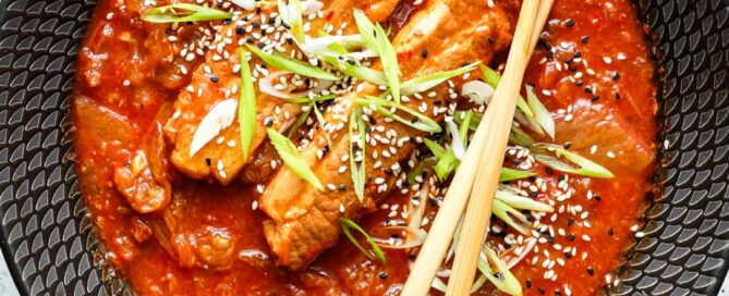 A large black bowl of Kimchi Pork Stew scattered with sesame seeds, nigella seeds and spring onion with chopsticks on the side.