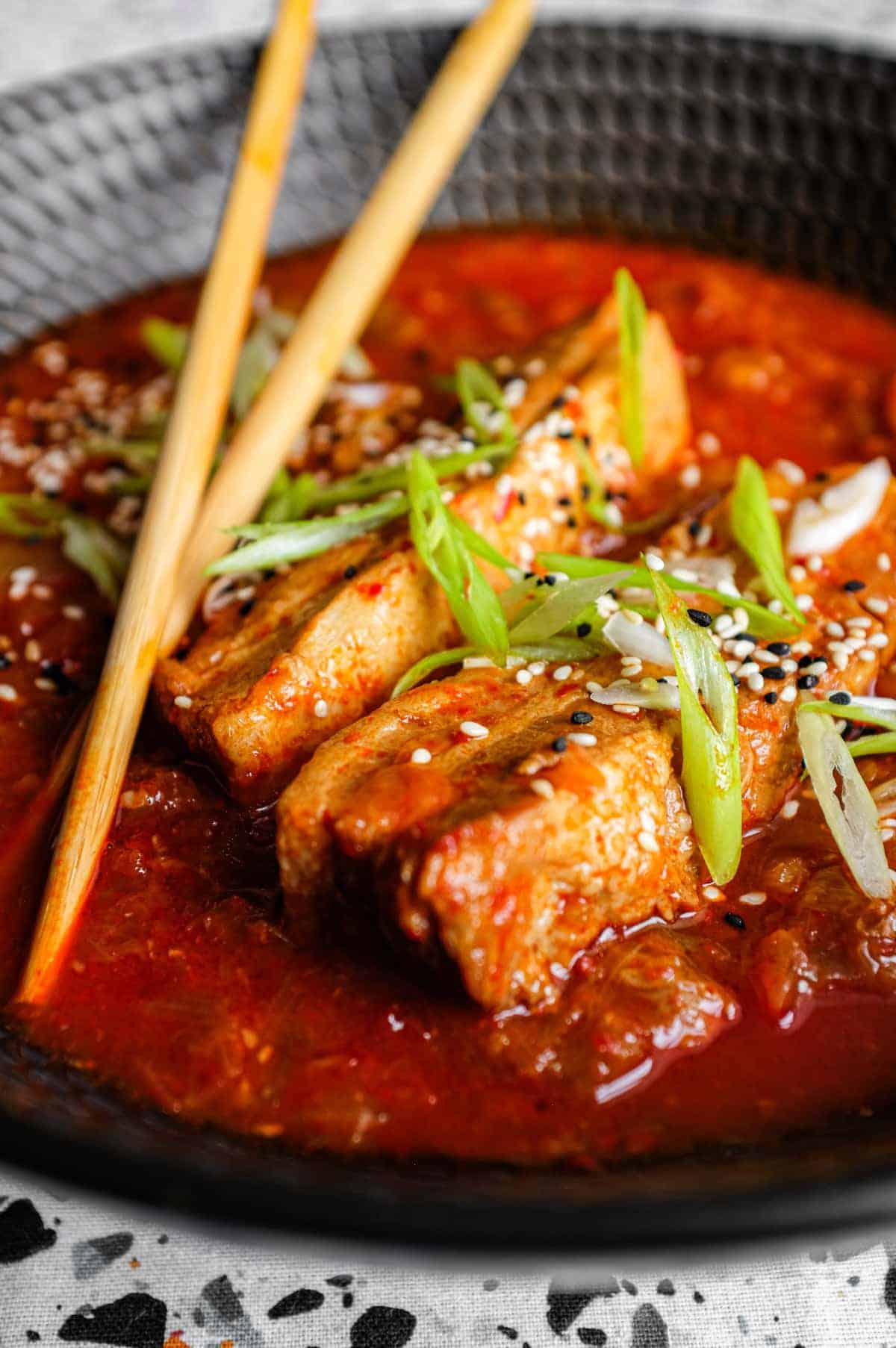 A large black bowl of Kimchi Pork Stew scattered with sesame seeds, nigella seeds and spring onion with chopsticks on the side.