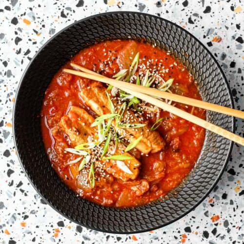 A large black bowl of Kimchi Pork Stew scattered with sesame seeds, nigella seeds and spring onion with chopsticks on the side.