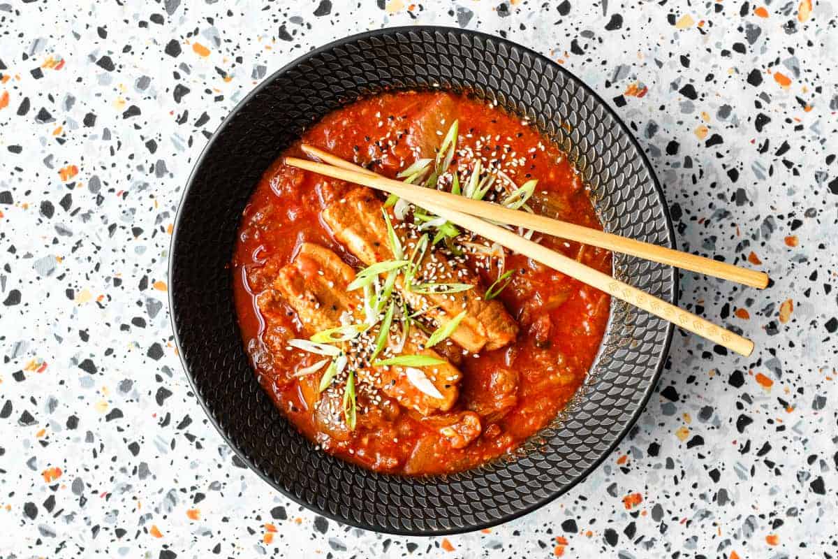 A large black bowl of Kimchi Pork Stew scattered with sesame seeds, nigella seeds and spring onion with chopsticks on the side.