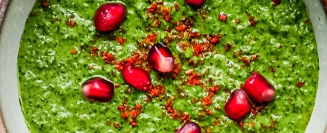 A small bowl of Zhoug Save with a spoon. Garnished with red pepper flakes and fresh pomegranate seeds.