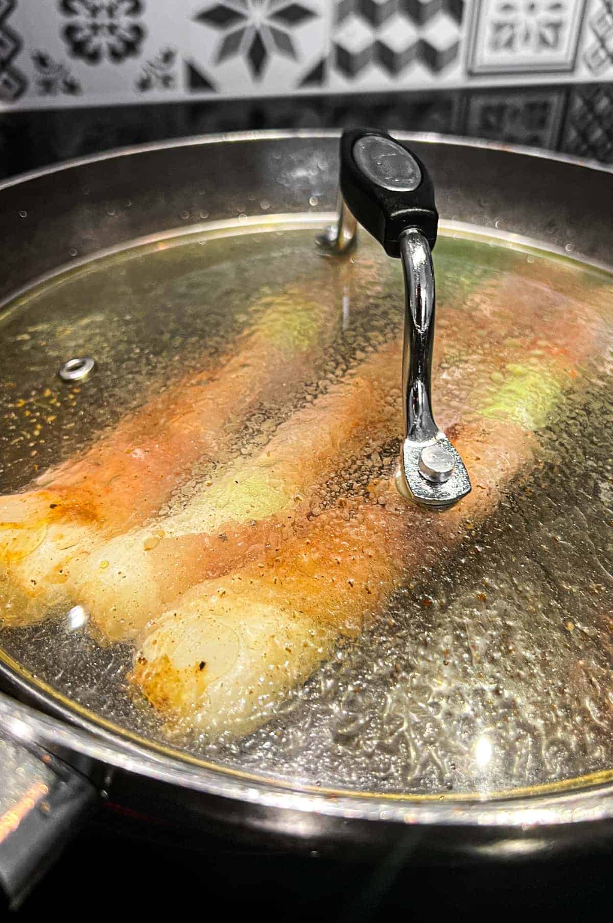 Spring onions wrapped in pancetta in a frying pan. A lid sits over the pan to steam the onions before frying until crisp.