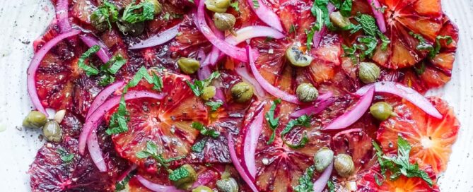 A large platter of thinly sliced blood oranges garnished with capers, onion and mint.