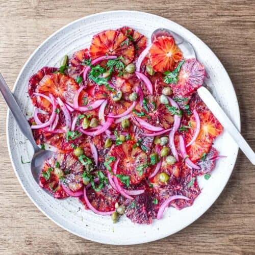 A large platter of thinly sliced blood oranges garnished with capers, onion and mint.