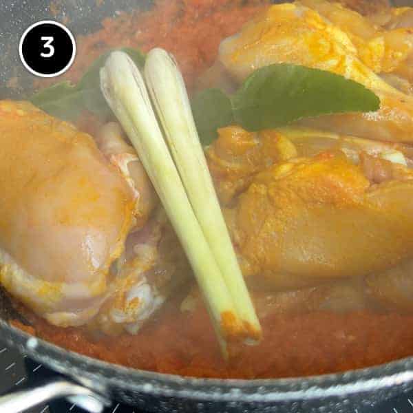 chicken cooking in pan with a chilli paste. On top are 3 lemongrass stalks and some kaffir lime leaves.