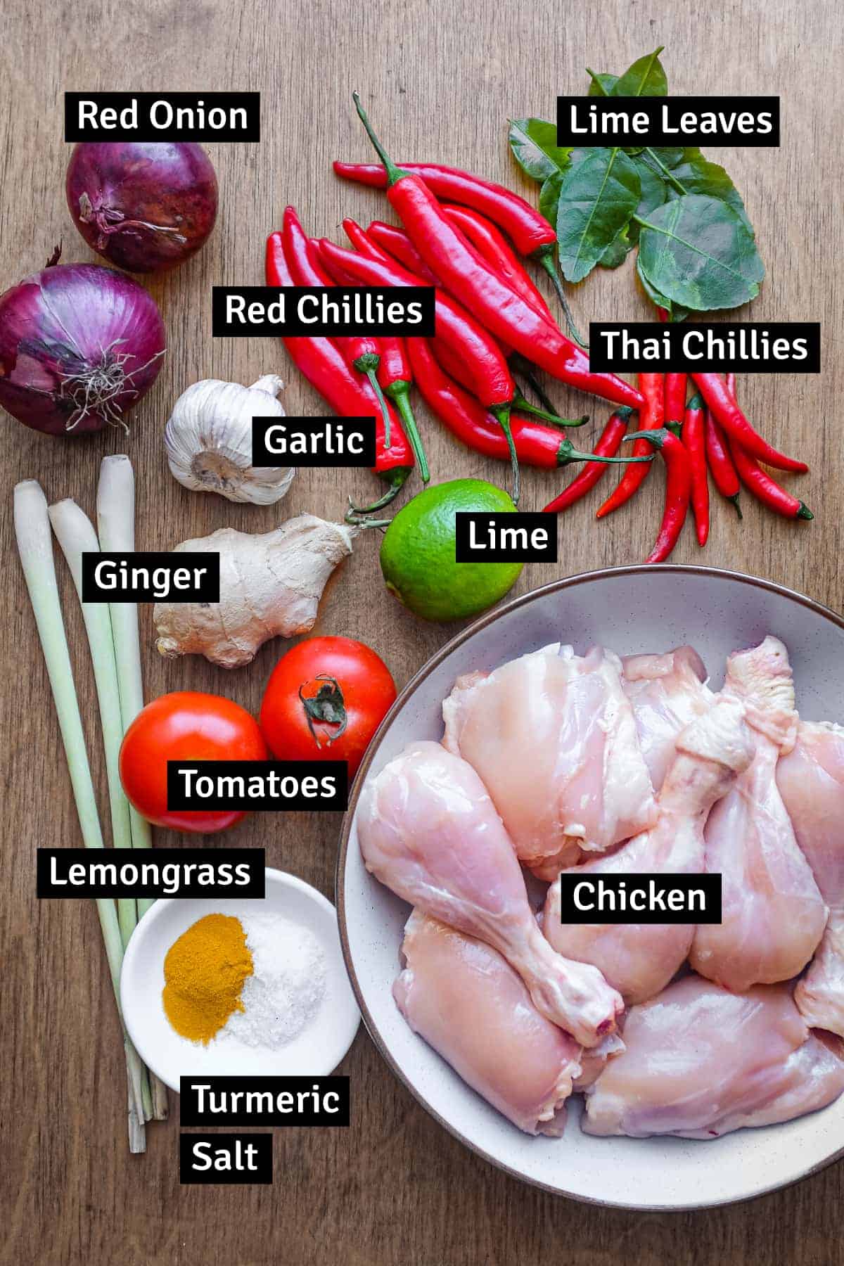 The ingredients for Ayam Rica Rica are arranged on a wooden surface: Chicken pieces, tomato, chillies, ginger, garlic, salt, turmeric powder, lime, lemongrass, onions and kaffir lime leaves.