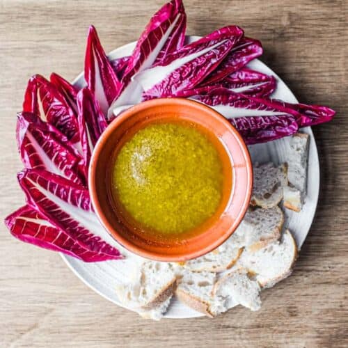 A small heated bowl of Italian Garlic & Anchovy sauce (Bagna Cauda). Radichhio leaves and small pieces of bread are arranged around the smallwer bowl on a larger platter.