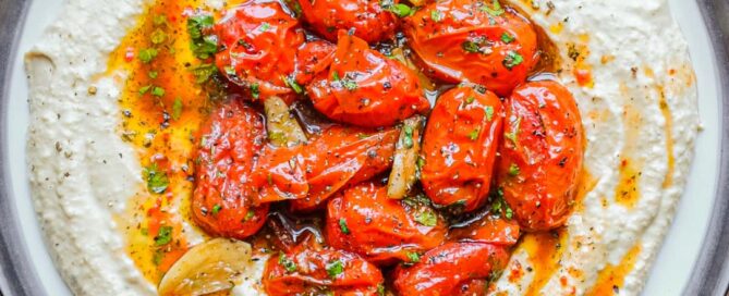 A platter of hummus with a centre filled with roasted tomatoes, garnished with pepper and herbs.