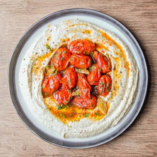 A platter of hummus with a centre filled with roasted tomatoes, garnished with pepper and herbs.