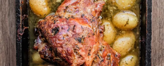 A roasting tin holding a roast lamb leg. Potatoes sit in the juices from the lamb.