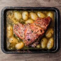 A roasting tin holding a roast lamb leg. Potatoes sit in the juices from the lamb.