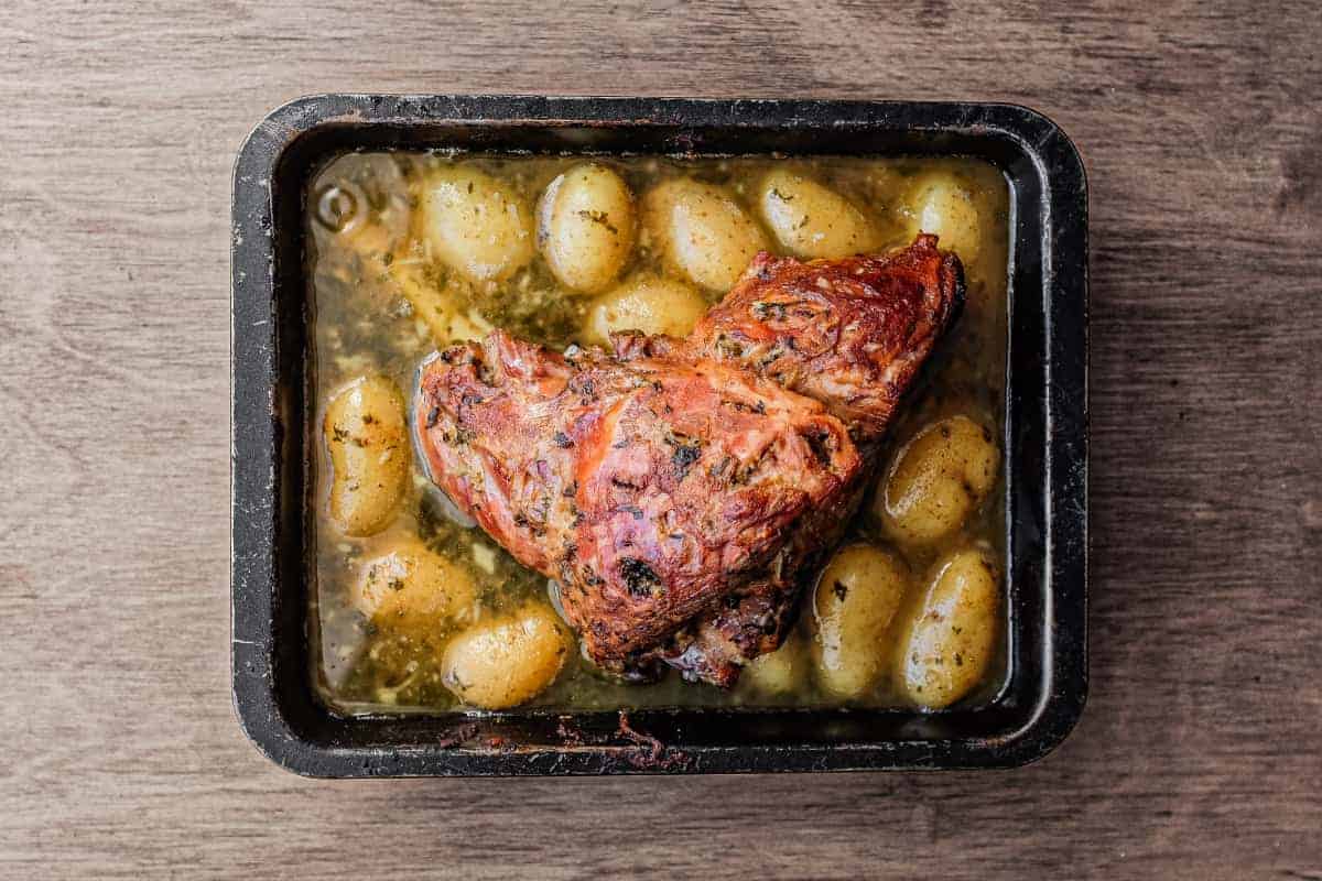 A roasting tin holding a roast lamb leg. Potatoes sit in the juices from the lamb.