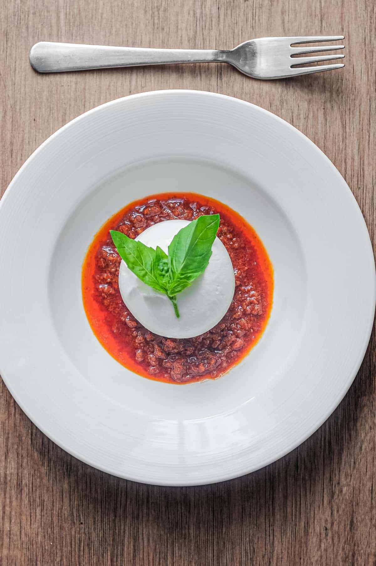 A plate of nduja sausage in liquid form, topped with a ball of burrata cheese and garnished with a small sprig of basil. A fork sits alongside.