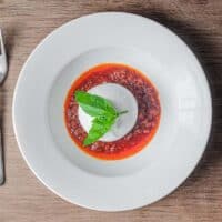 A plate of nduja sausage in liquid form, topped with a ball of burrata cheese and garnished with a small sprig of basil. A fork sits to the left.