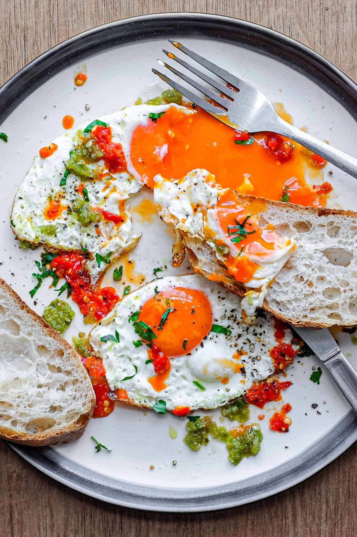 A plate with two fried eggs dotted with spicy Shatta chilli sauce. One of the yolks has been burst and the yelllow yolk oozes onto the plate.