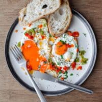 A plate with two fried eggs dotted with spicy Shatta chilli sauce. One of the yolks has been burst and the yelllow yolk oozes onto the plate.