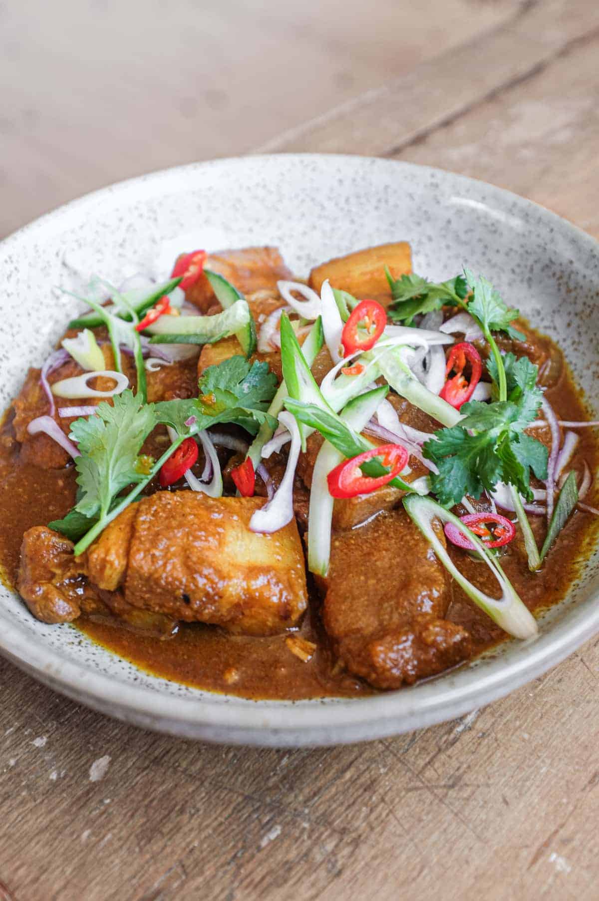 A bowl of Gaeng Hung Lay (Thai Pork Curry) garnished with herbs, spring onion, chilli and shallots