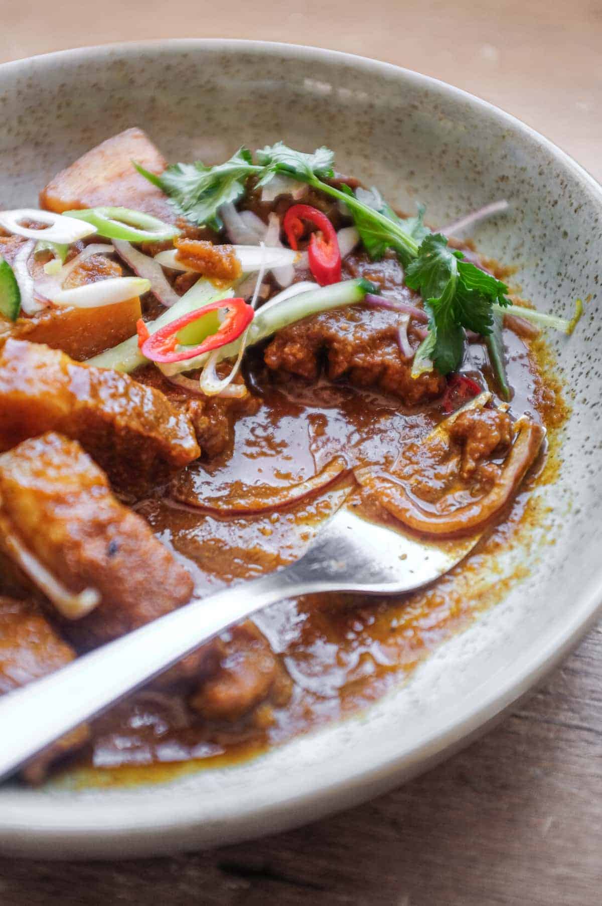 A bowl of Gaeng Hung Lay (Thai Pork Curry) garnished with herbs, spring onion, chilli and shallots