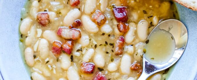 A bowl of French garlic soup with beans and bacon. A spoon sits in the bowl alongside a slice of crusty bead.