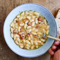 A bowl of French garlic soup with beans and bacon. A spoon sits in the bowl alongside a slice of crusty bead.