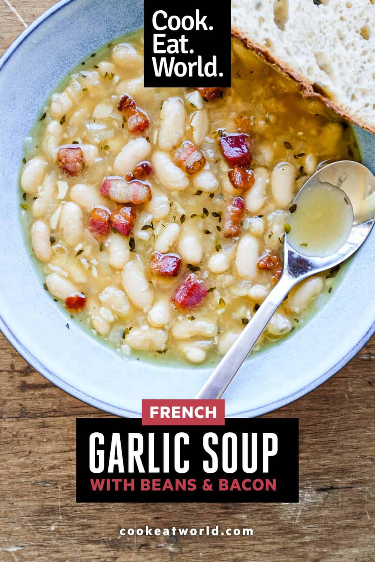 A bowl of French garlic soup with beans and bacon. A spoon sits in the bowl alongside a slice of crusty bead.