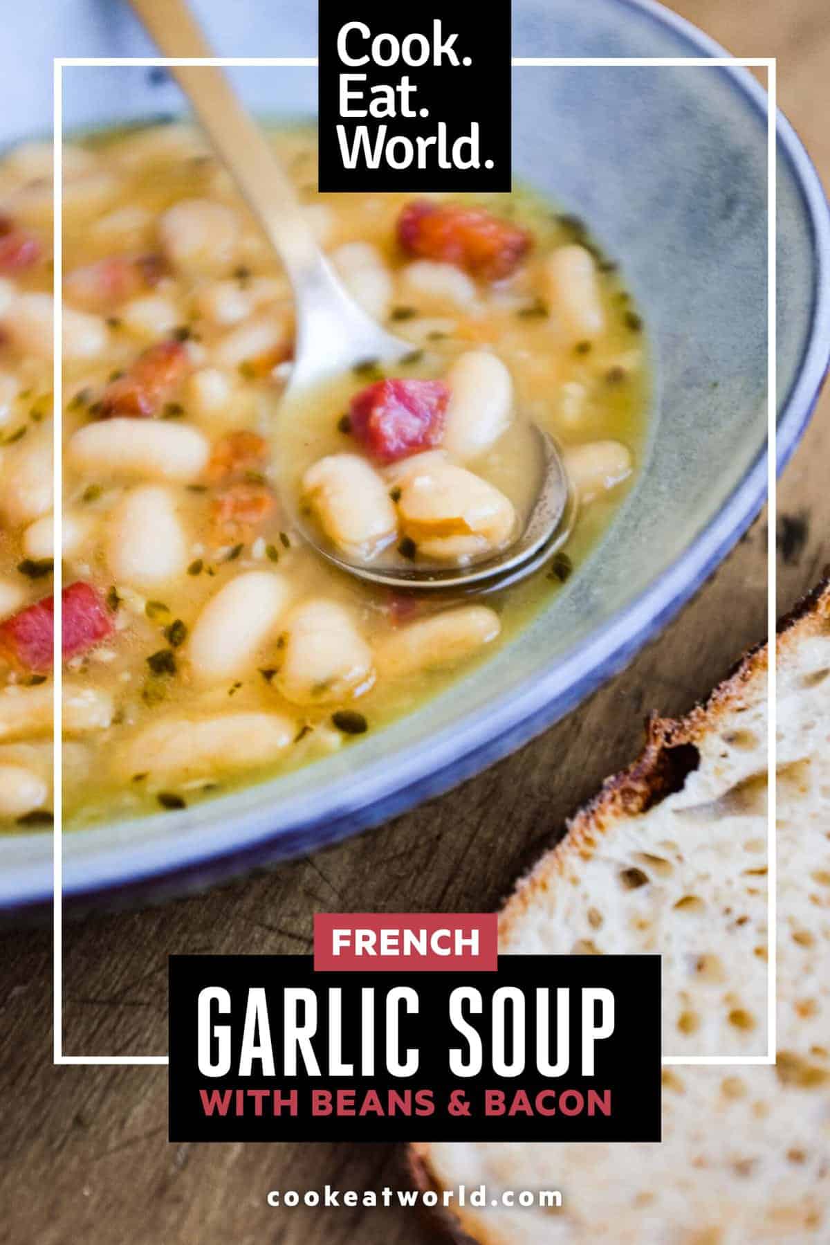 A bowl of French garlic soup with beans and bacon. A spoon sits in the bowl alongside a slice of crusty bead.