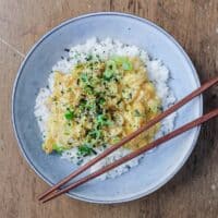 A bowl of Japanese scrambled eggs over rice and garnished with rice seasoning and spring onions. Wooden chopsticks sit on the bowl.