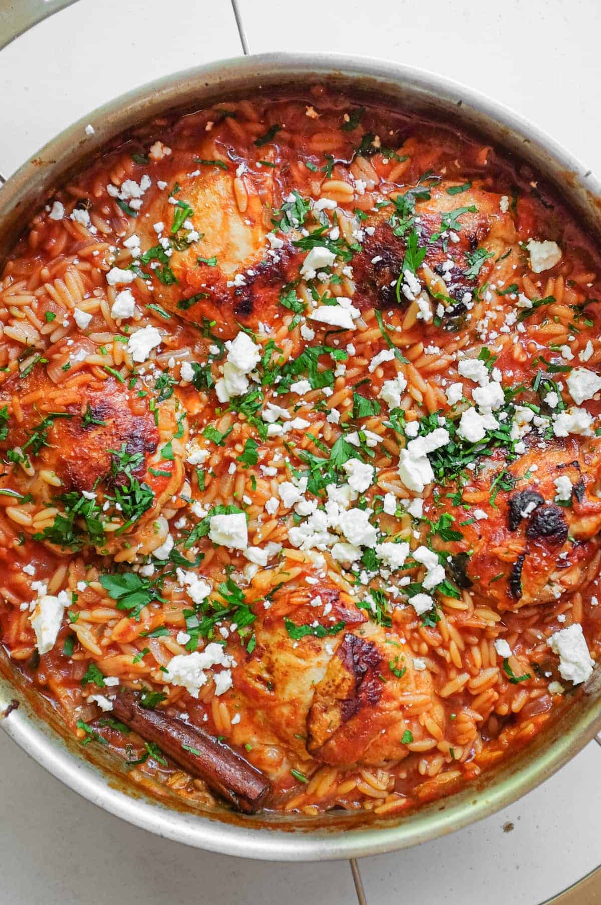 A pan of Youvetsi Chicken is garnished with fresh parsley and feta cheese.