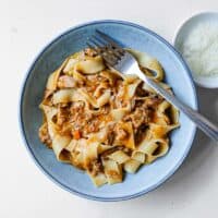A bowl of duck ragù tossed in pappardelle pasta sits with a small bowl of Parmigiano Reggiano cheese