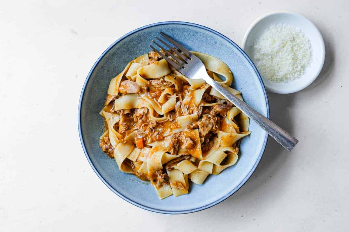 A bowl of duck ragù tossed in pappardelle pasta sits with a small bowl of Parmigiano Reggiano cheese