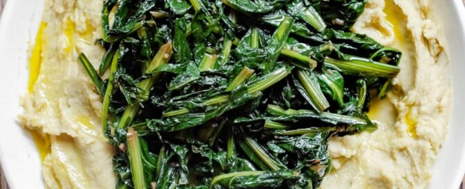 An overhead photograph of a large platter of fava bean puree topped with dark green chicory leaves