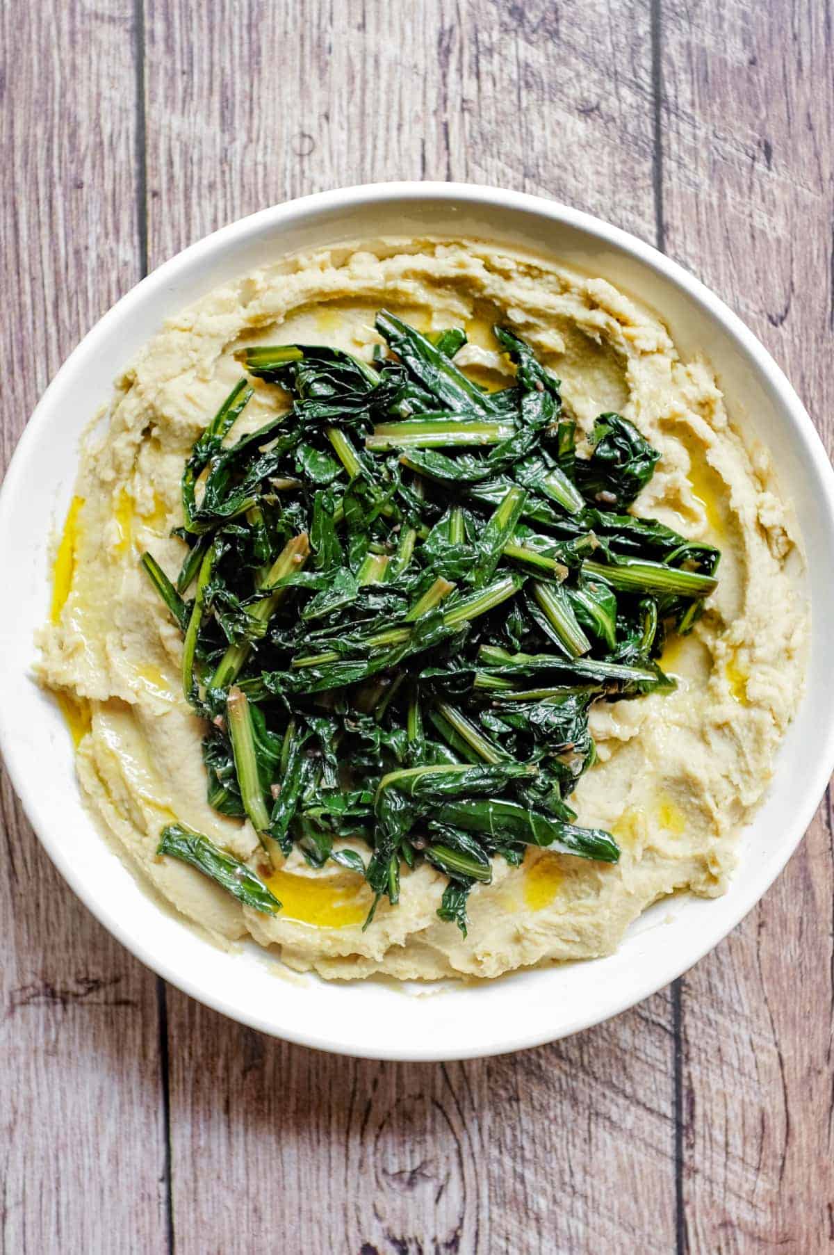 An overhead photograph of a large platter of fava bean puree topped with dark green chicory leaves