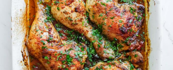 Four chicken quarters sit in a vibrant red harissa sauce surrounded by black olives and tomatoes. The dish is scattered with fresh finely chopped parsley.