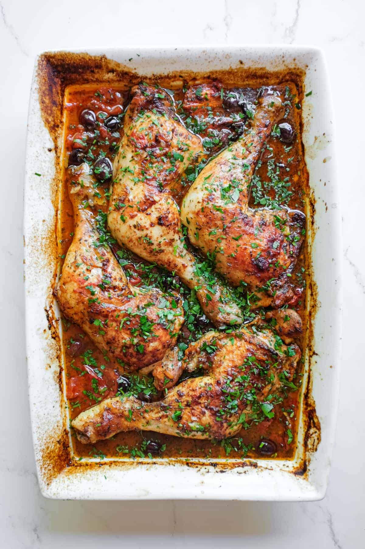 Four chicken quarters sit in a vibrant red harissa sauce surrounded by black olives and tomatoes. The dish is scattered with fresh finely chopped parsley.