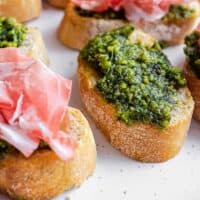 A serving platter of toasted bread with homemade pistachio pesto, some topped with cured ham.