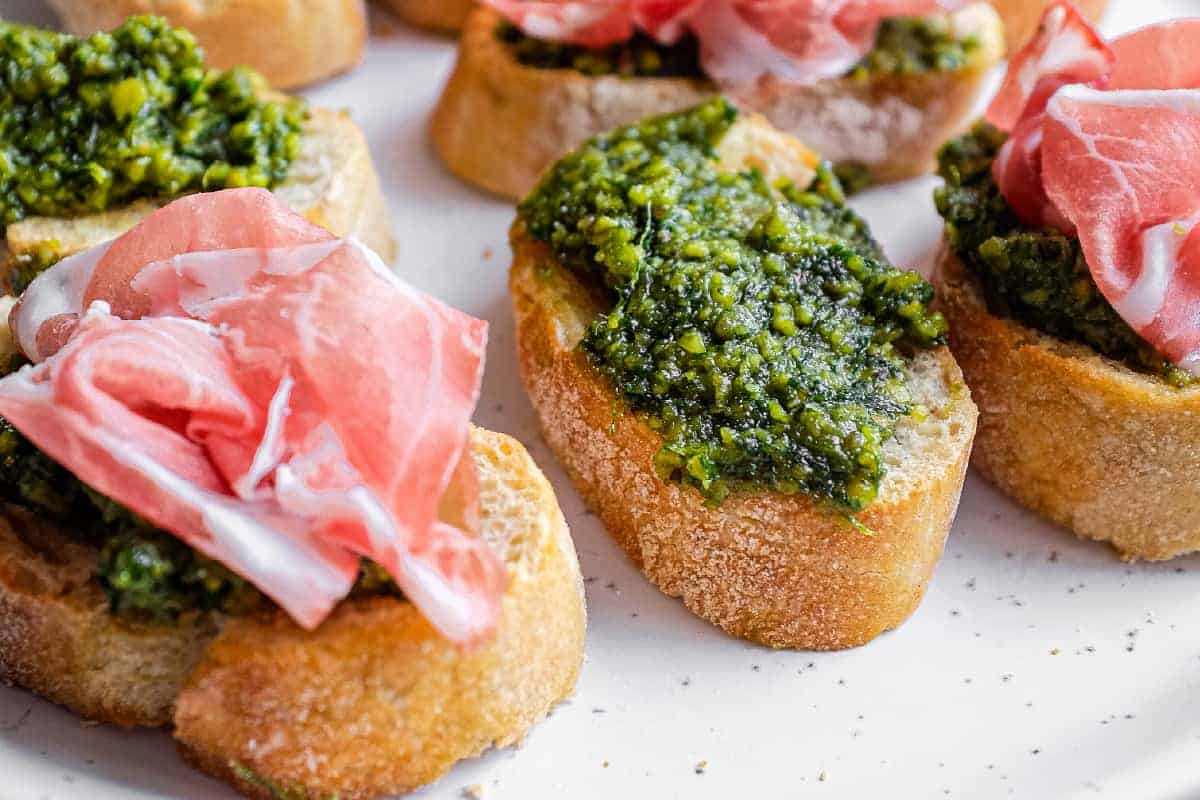 A serving platter of toasted bread with homemade pistachio pesto, some topped with cured ham.