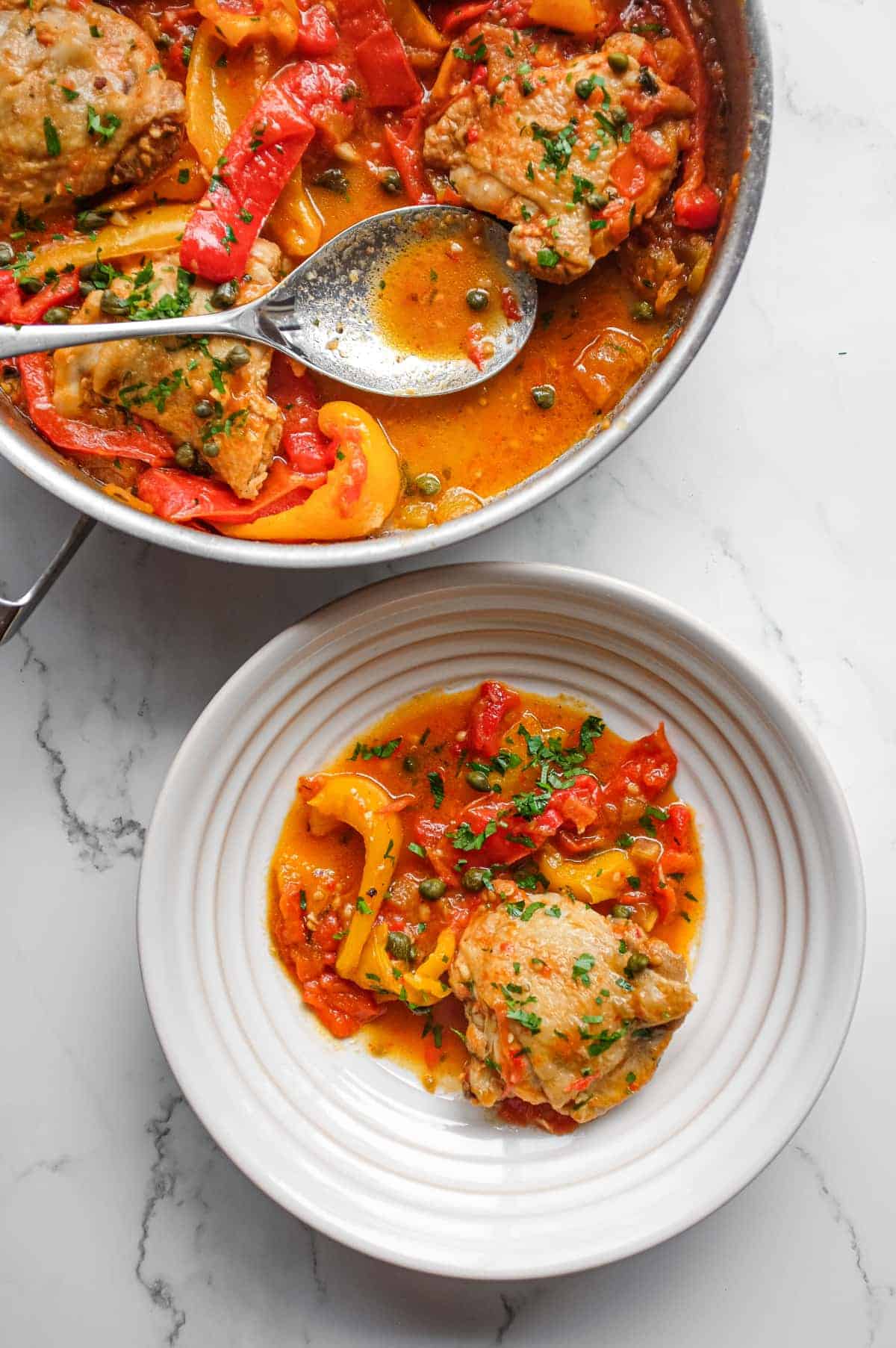 A pan of Roman chicken stew with tender chicken, sweet peppers, capers and chopped parsley alongside a portion in a small white bowl.