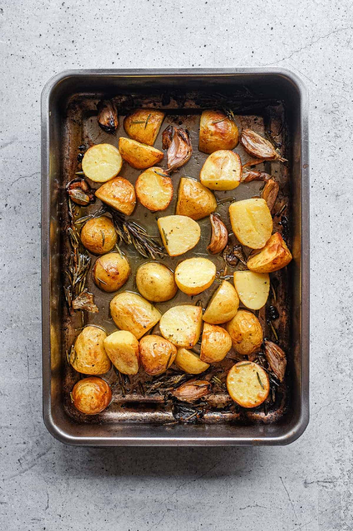 Tender roast potato pieces coated with olive oil, rosemary and garlic in a baking tin.